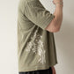 The right side of someone wearing a yellow brownish green t-shirt with a cream illustration of tong ho. His hand is raised close to his face.