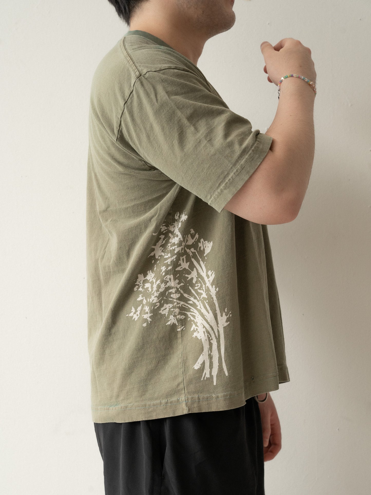 The right side of someone wearing a yellow brownish green t-shirt with a cream illustration of tong ho. His hand is raised close to his face.