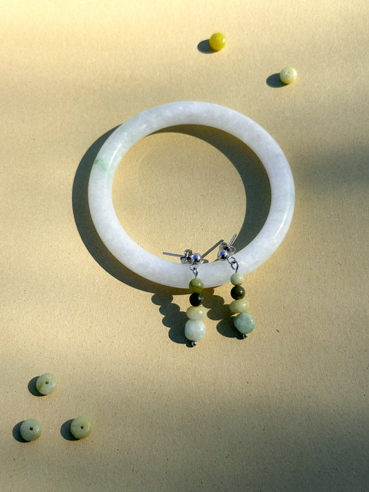 A pair of green earrings made of jade, moonstone, and stainless steel hang on a jade bracelet that sits on top of a brownish green background next to individual gemstones.
