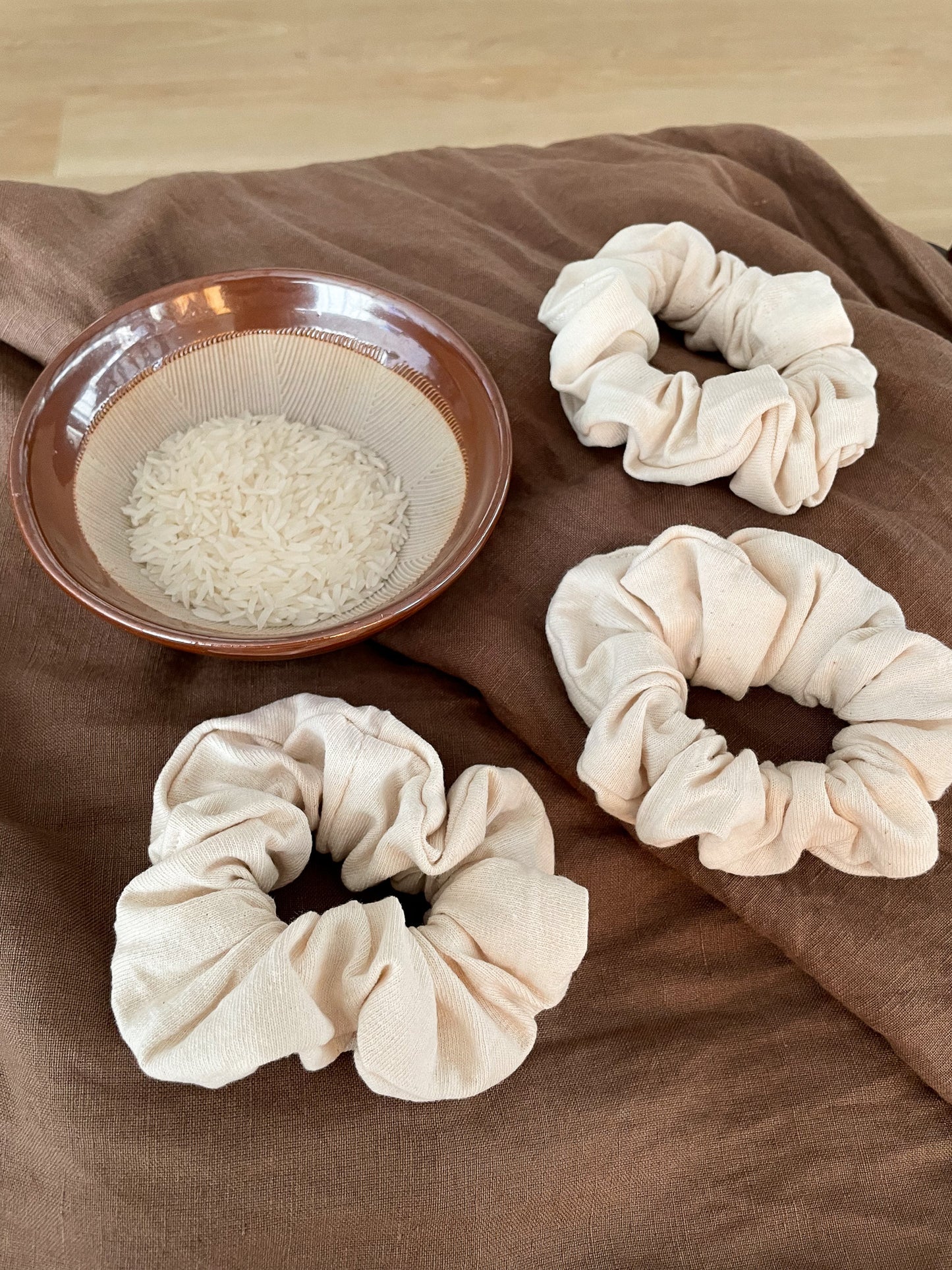 Three off white scrunchies lay flat on a brown cloth next to a ceramic bowl of uncooked rice.