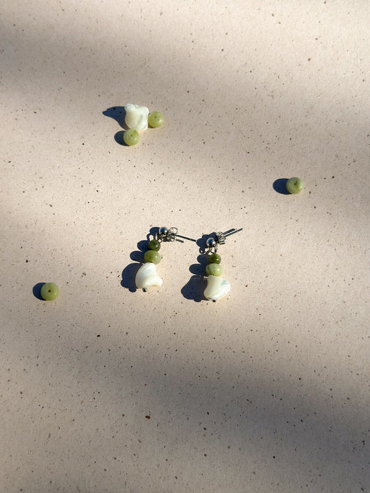 A pair of white and green earrings made of jade, pearl, and stainless steel on a cream speckled background next to individual gemstones and pearls.