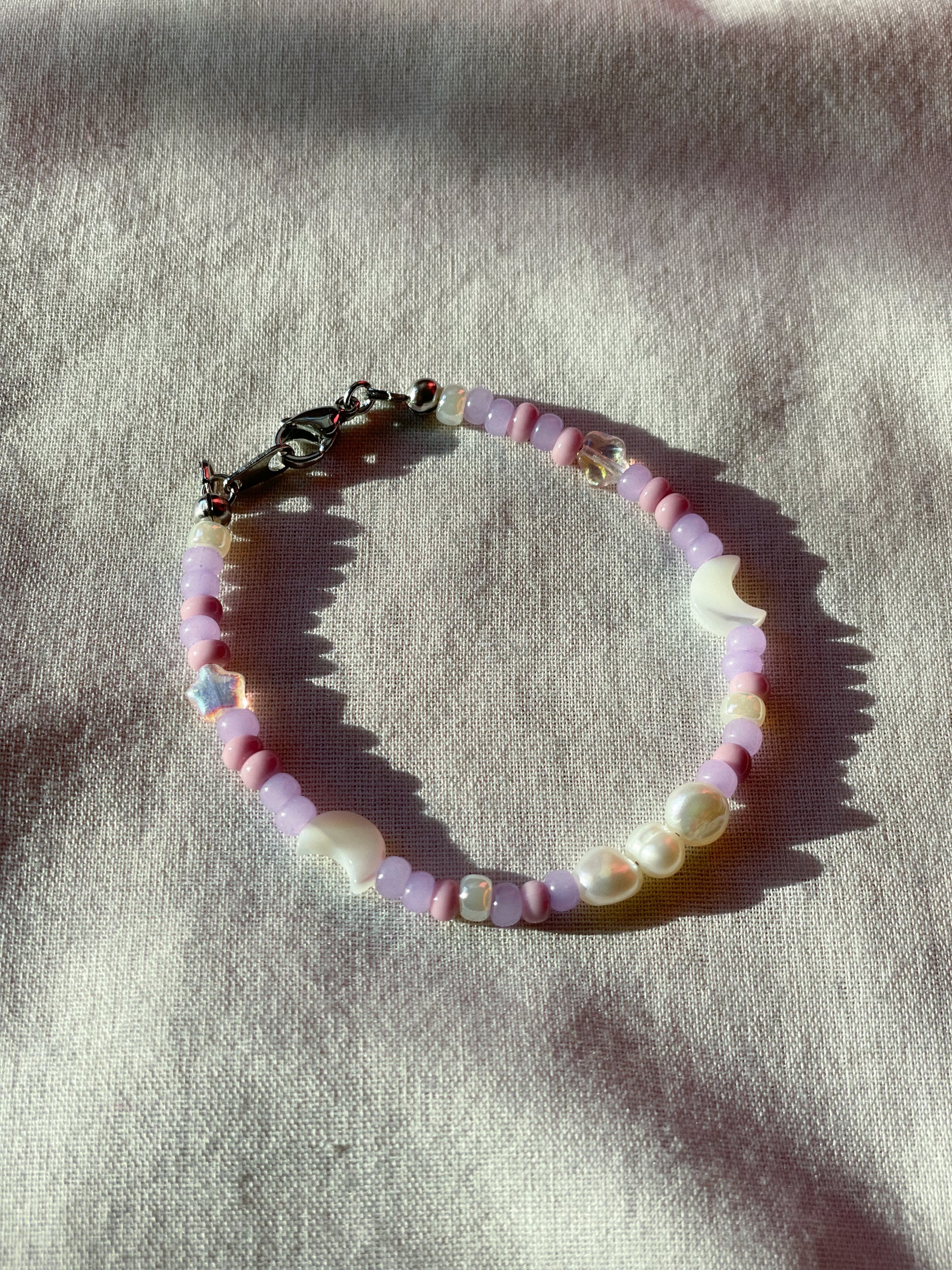 A bracelet made of purple Czech glass beads, star-shaped Czech glass beads, purple freshwater pearls, moon-shaped white freshwater pearls, and stainless steel lay flat on a white piece of cloth.