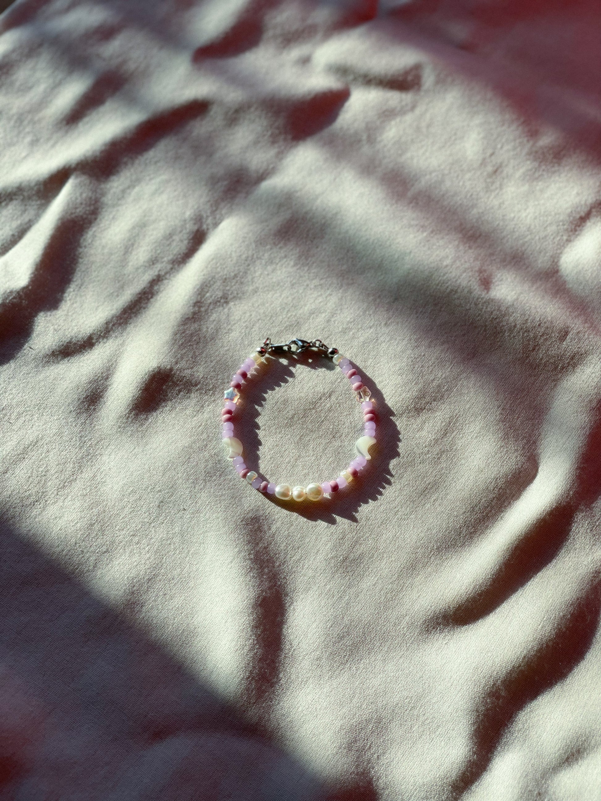 A bracelet made of purple Czech glass beads, star-shaped Czech glass beads, purple freshwater pearls, moon-shaped white freshwater pearls, and stainless steel lay flat on a white piece of cloth.