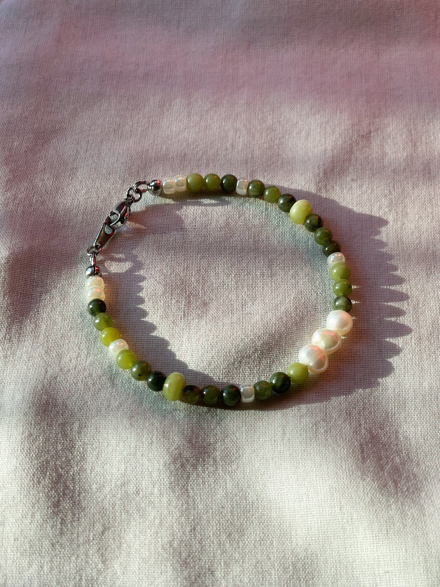 A bracelet made of jade, Japanese glass beads, white freshwater pearls, and stainless steel lay flat on a white piece of cloth.