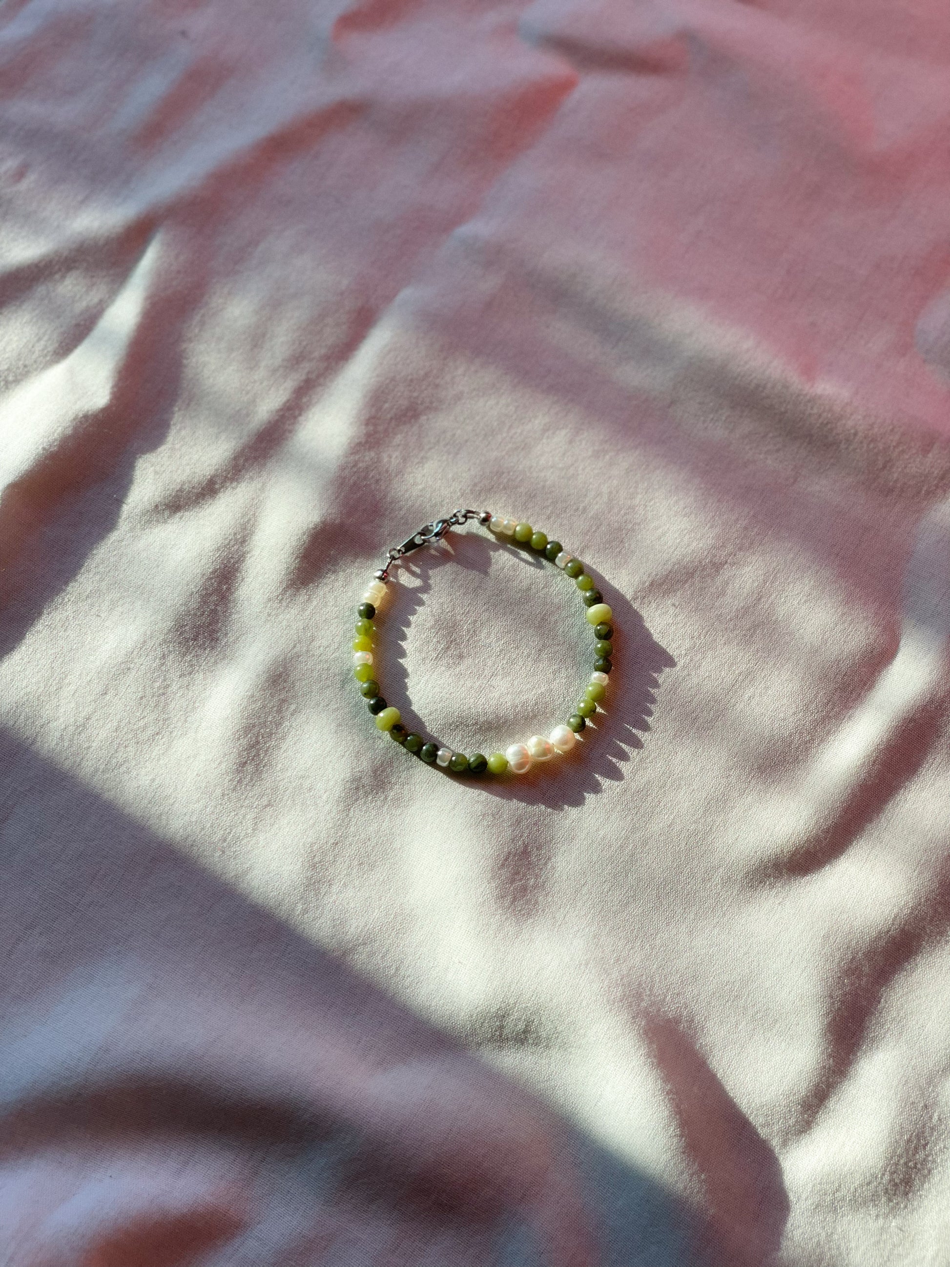 A bracelet made of jade, Japanese glass beads, white freshwater pearls, and stainless steel lay flat on a white piece of cloth.