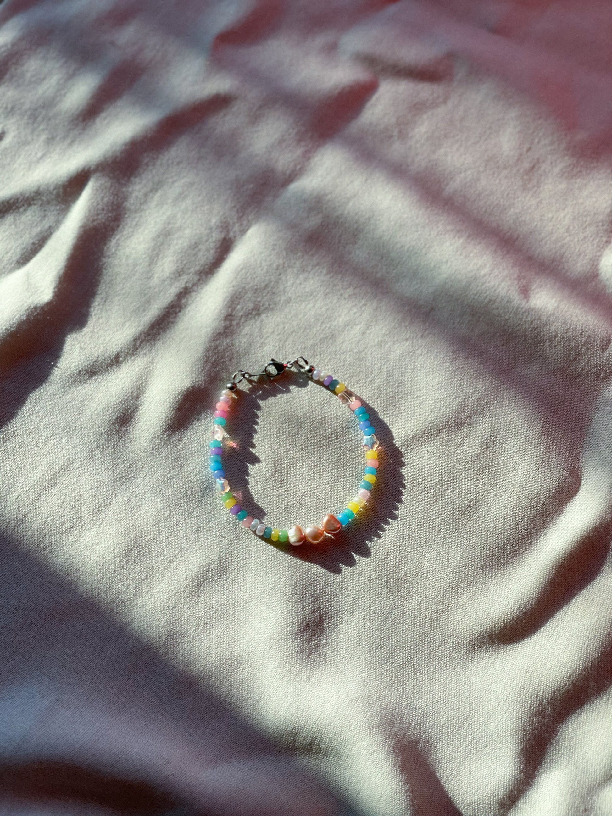 A bracelet made of round colorful Czech glass beads, star-shaped Czech glass beads, purple freshwater pearls, and stainless steel lay flat on a white piece of cloth.