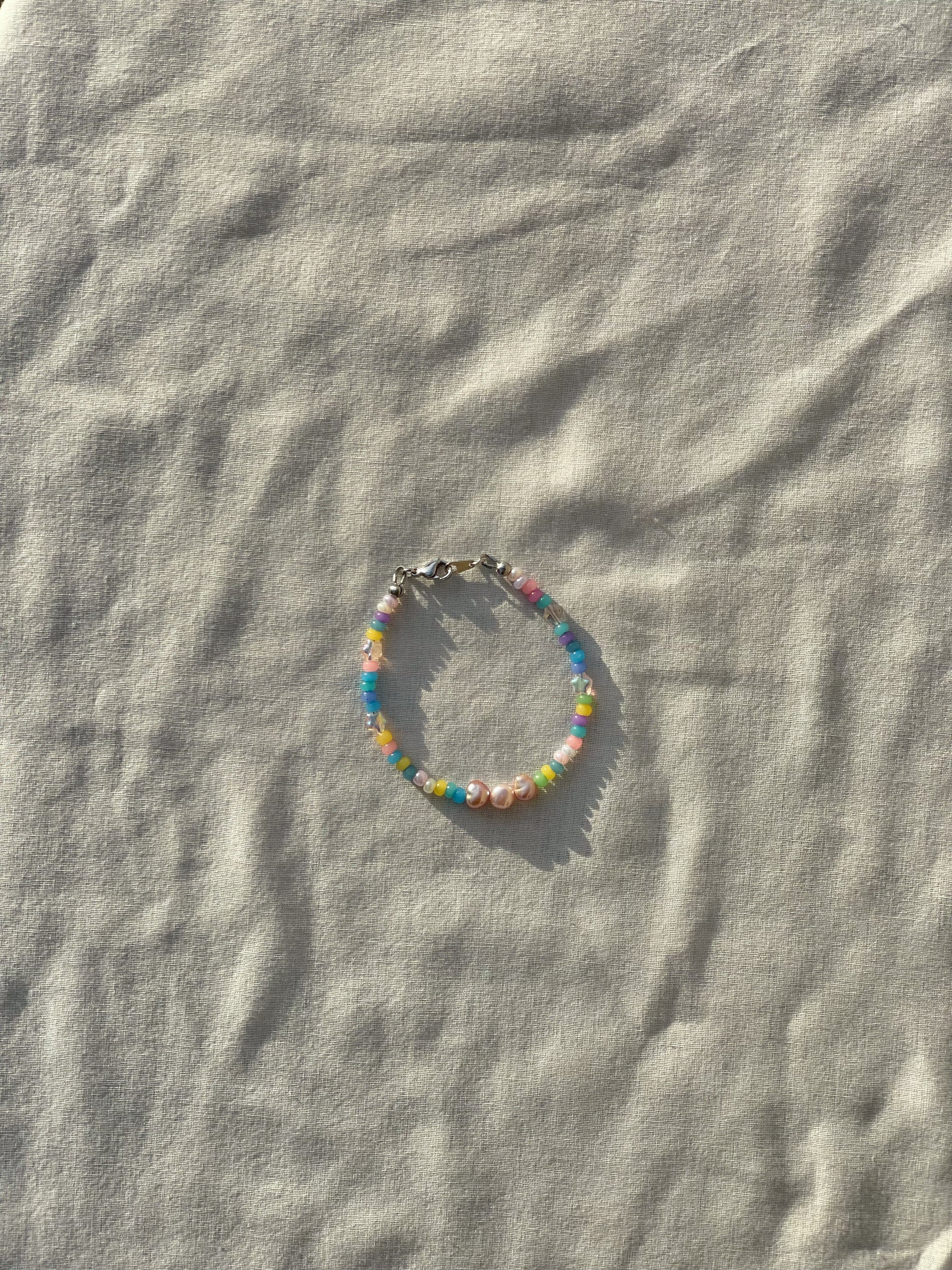 A bracelet made of round colorful Czech glass beads, star-shaped Czech glass beads, purple freshwater pearls, and stainless steel lay flat on a white piece of cloth in the sunlight.