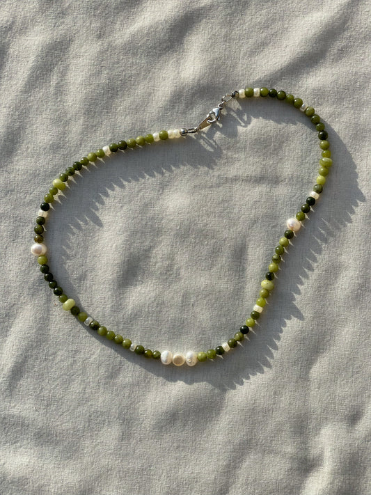 A necklace made of jade, Japanese glass beads, white freshwater pearls, and stainless steel lay flat on a white piece of cloth in the sunlight.