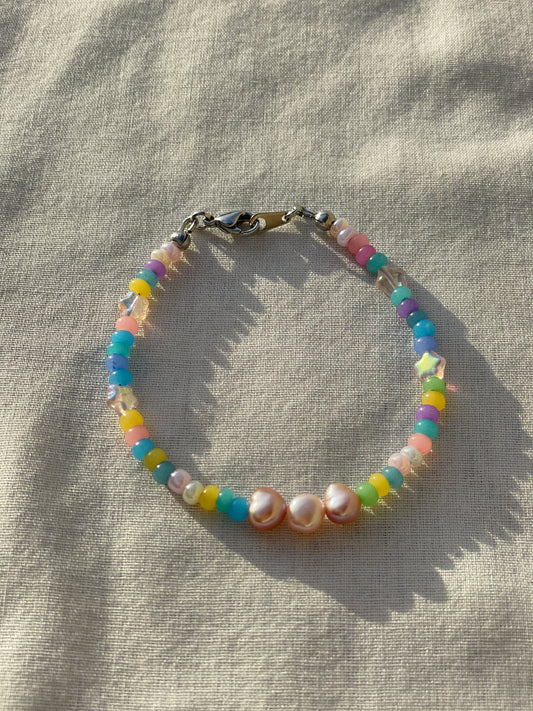 A bracelet made of round colorful Czech glass beads, star-shaped Czech glass beads, purple freshwater pearls, and stainless steel lay flat on a white piece of cloth in the sunlight.