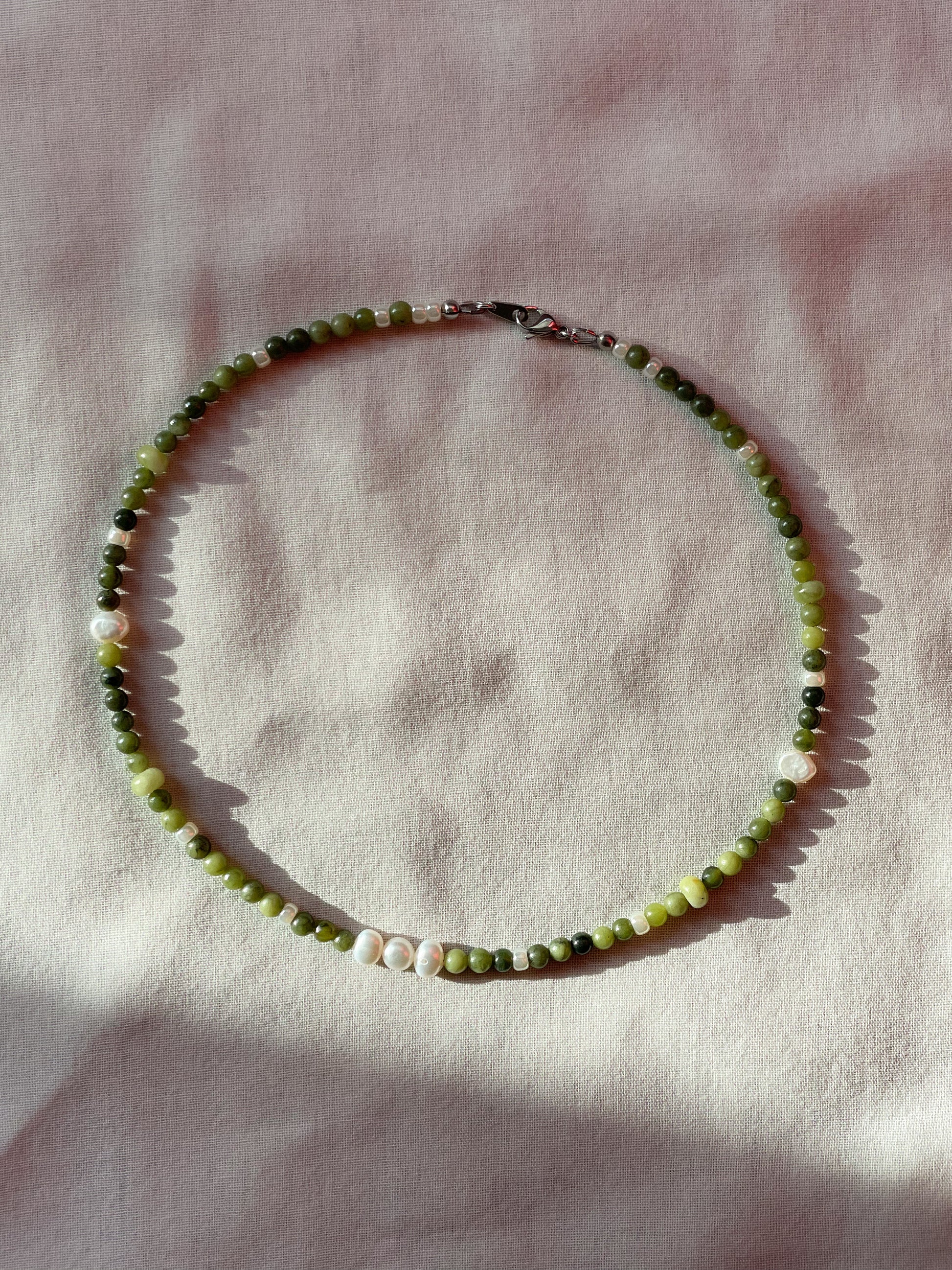 A necklace made of jade, Japanese glass beads, white freshwater pearls, and stainless steel lay flat on a white piece of cloth.