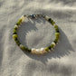 A bracelet made of jade, Japanese glass beads, white freshwater pearls, and stainless steel lay flat on a white piece of cloth in the sunlight.