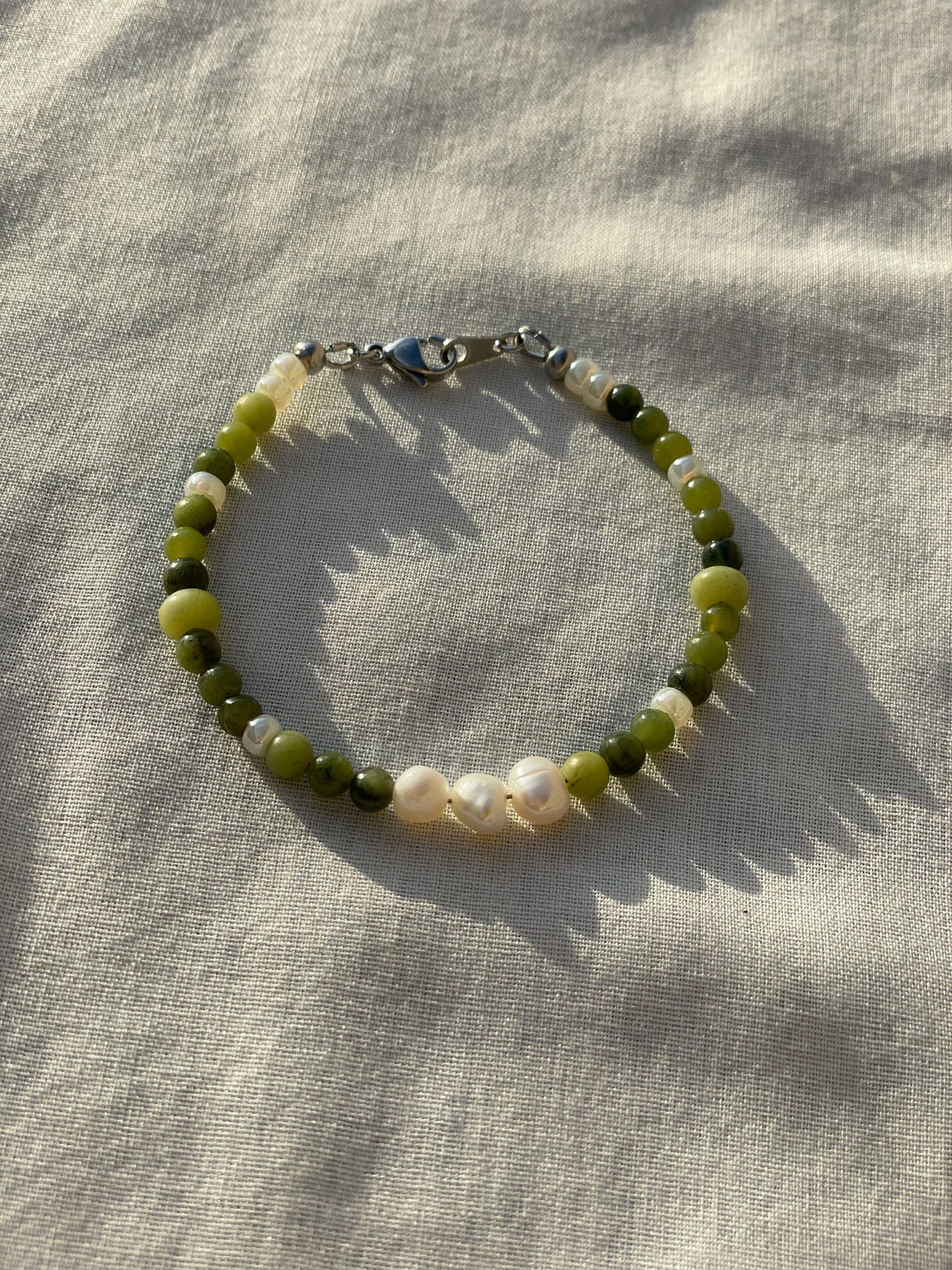 A bracelet made of jade, Japanese glass beads, white freshwater pearls, and stainless steel lay flat on a white piece of cloth in the sunlight.