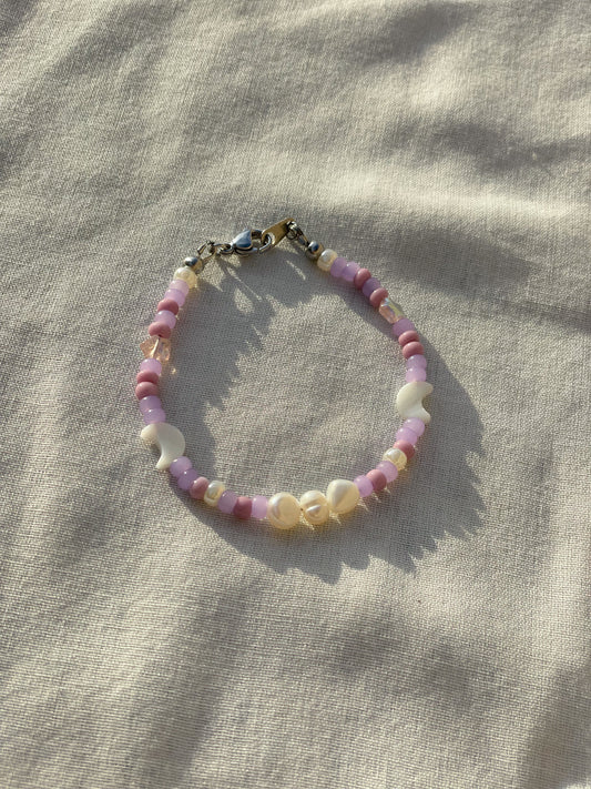 A bracelet made of purple Czech glass beads, star-shaped Czech glass beads, purple freshwater pearls, moon-shaped white freshwater pearls, and stainless steel lay flat on a white piece of cloth in the sunlight. 