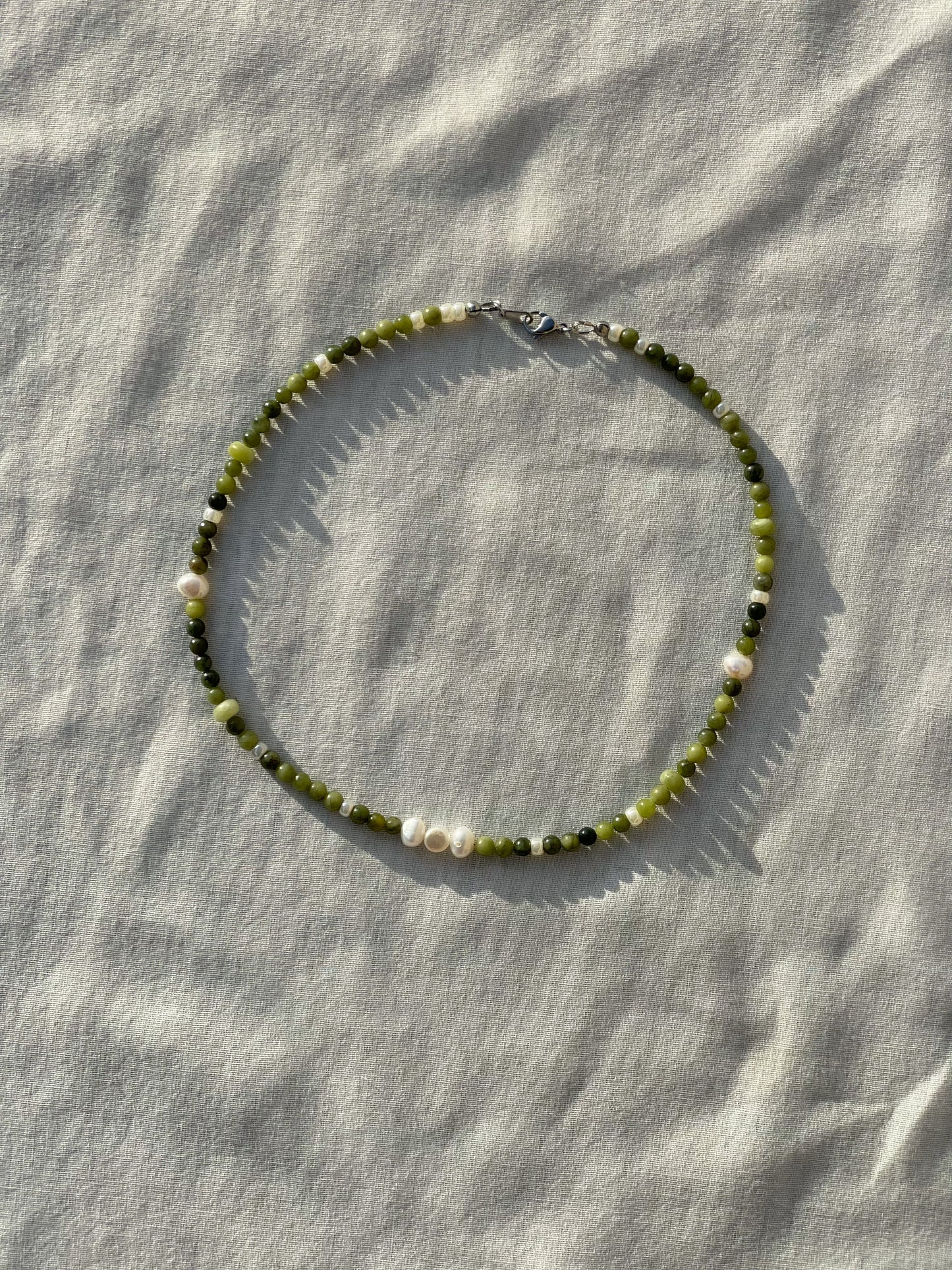 A necklace made of jade, Japanese glass beads, white freshwater pearls, and stainless steel lay flat on a white piece of cloth in the sunlight.