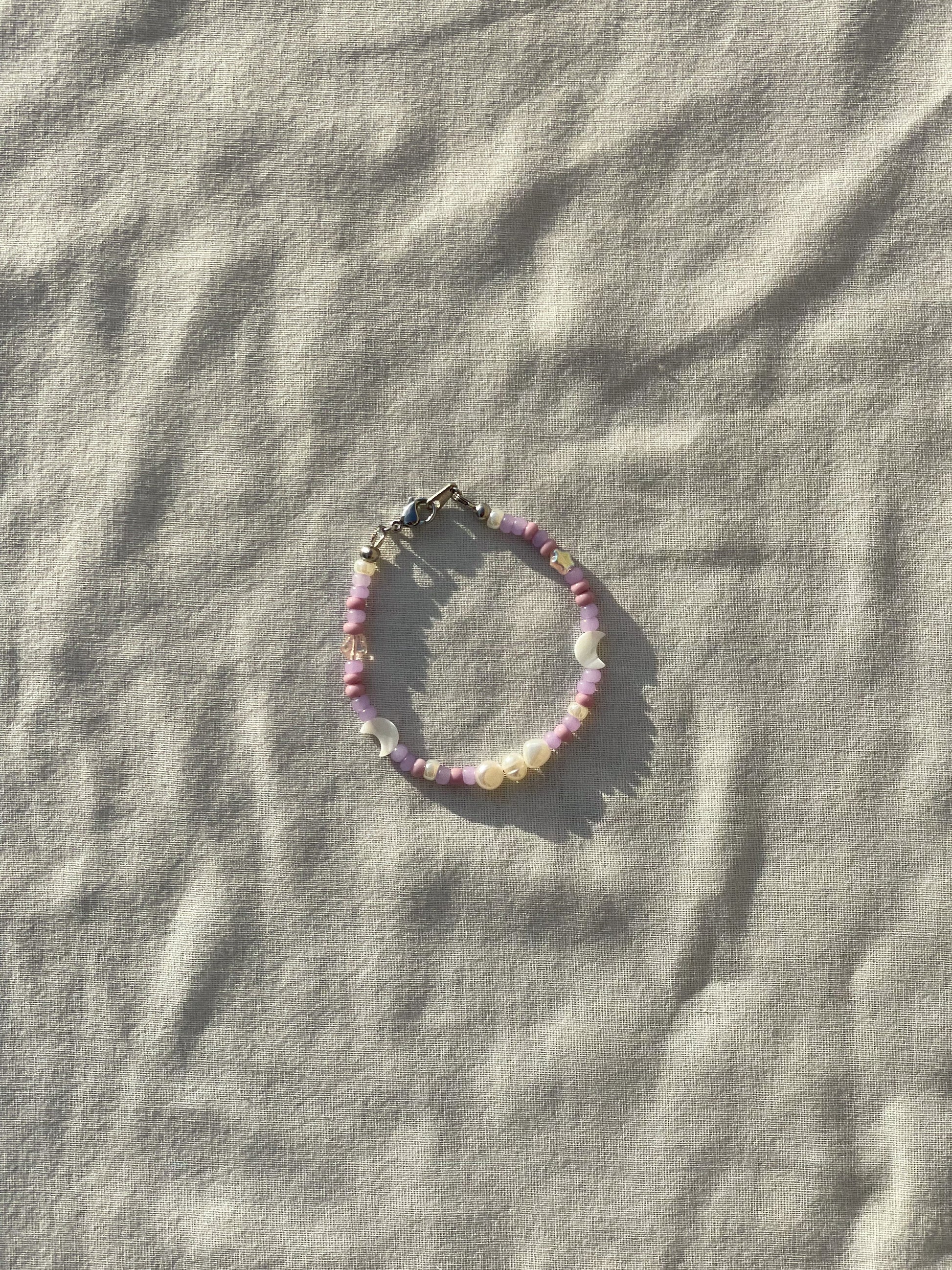 A bracelet made of purple Czech glass beads, star-shaped Czech glass beads, purple freshwater pearls, moon-shaped white freshwater pearls, and stainless steel lay flat on a white piece of cloth in the sunlight. 