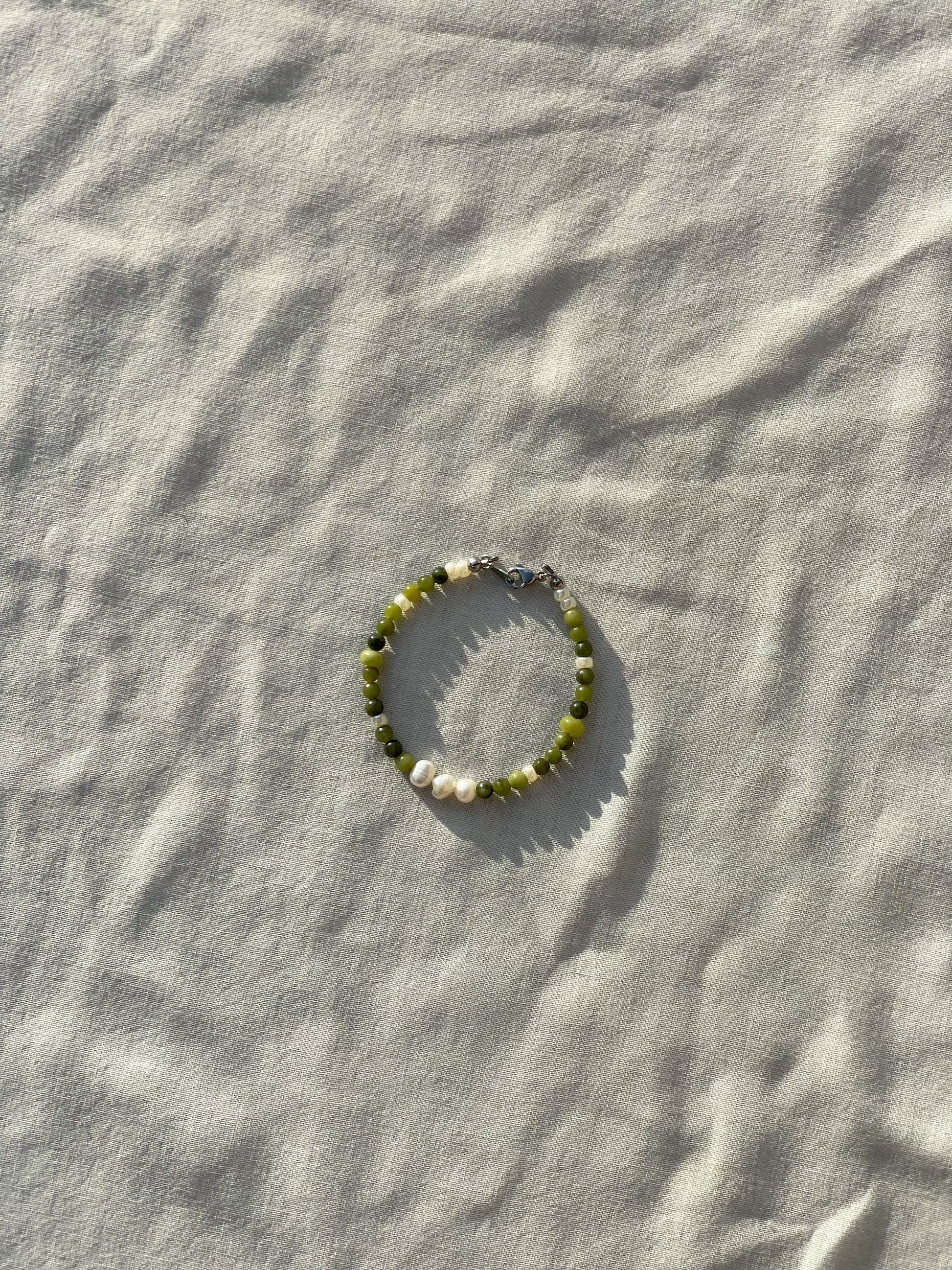 A bracelet made of jade, Japanese glass beads, white freshwater pearls, and stainless steel lay flat on a white piece of cloth in the sunlight.