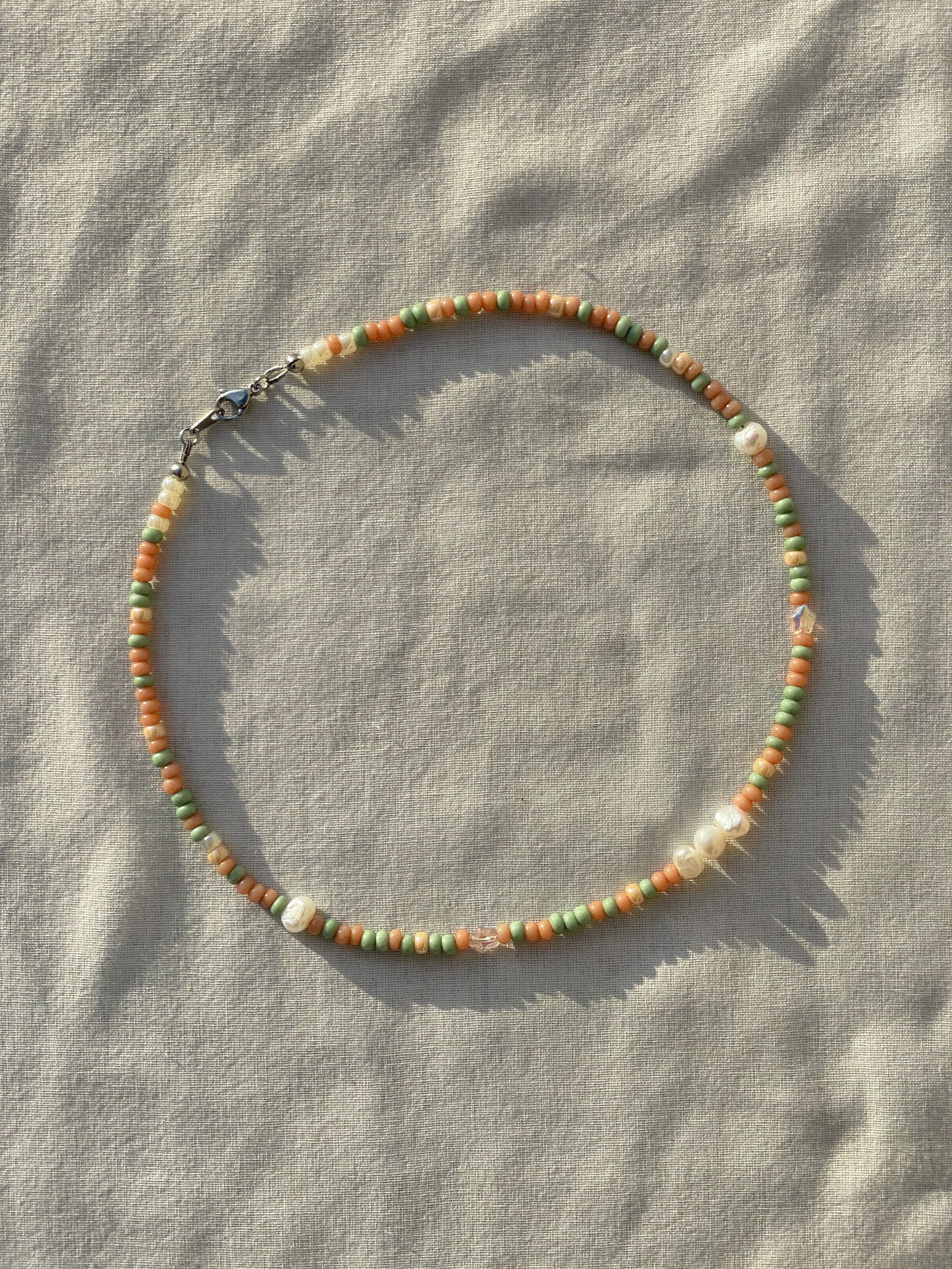 A necklace made of Czech glass beads, white freshwater pearls, and stainless steel lay flat on a white piece of cloth in the sunlight.